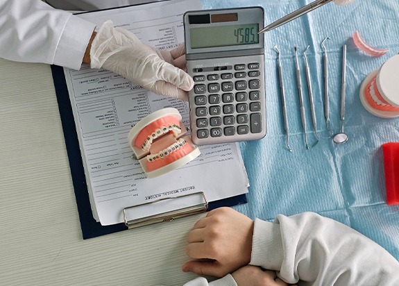 Bird’s eye view of gloved hand holding calculator out to patient next to model teeth with braces